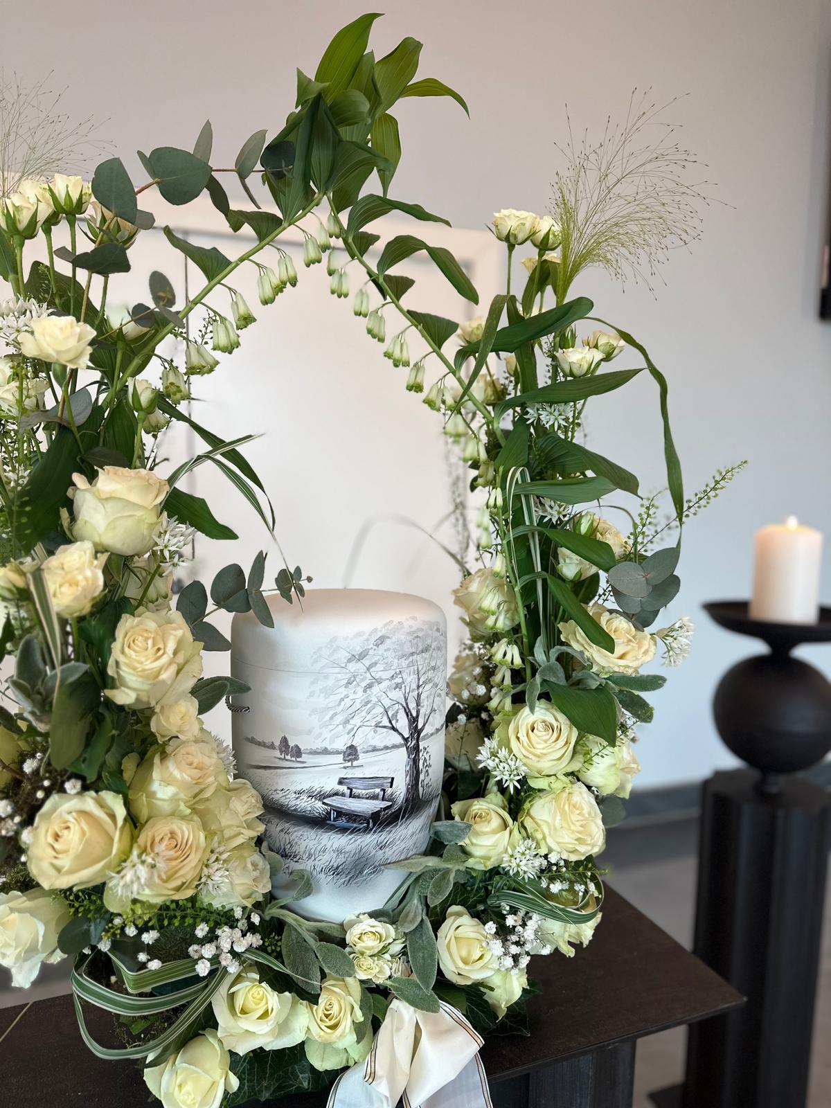 Urne mit Baum- und Bankmotiv, umrahmt von einem Blumenbogen aus weißen Rosen