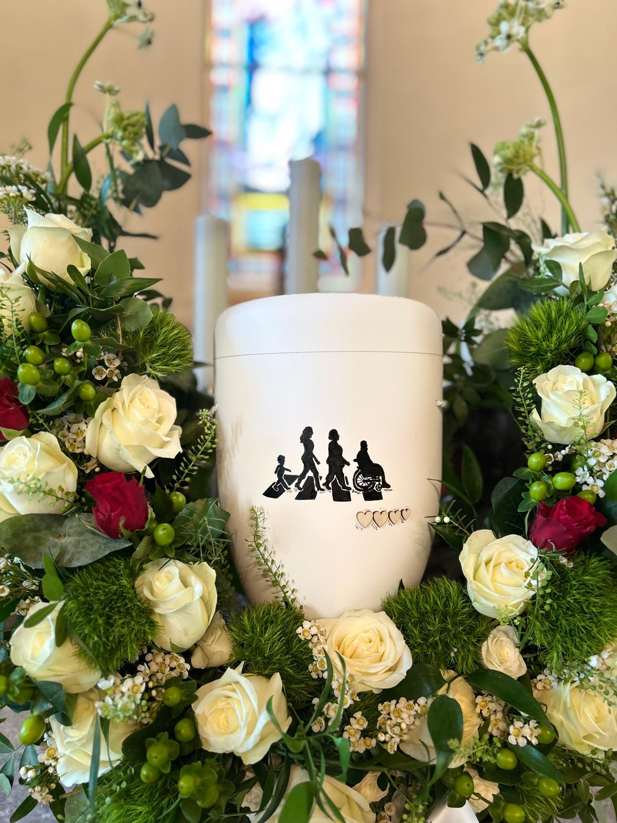 Weiße Urne mit Familien-Silhouette in einem Rosengesteck in der Kirche